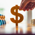 Hand holding wooden dollar sign between diverse people and coin stacks, symbolizing wealth distribution, income gap, economic decision-making, and social impact of financial inequality.